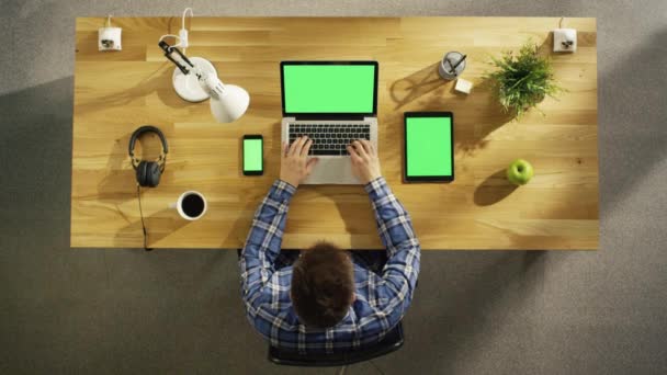 Pohled shora na kreativní mladý muž sedí na jeho stole psaní na notebooku vedle leží Smartphone a Tablet počítač, všech elektronických zařízení mají zelené maketa obrazovku. — Stock video
