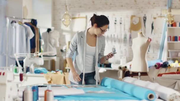 Hermosa morena diseñadora de moda establece plantillas en las telas y mira a su teléfono inteligente. Trabaja en un estudio de colores claros lleno de varias ropas, telas y bocetos en la pared . — Vídeos de Stock