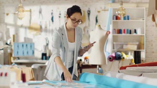 Bella Bruna Fashion Designer Stende i modelli sui tessuti e consulta il suo smartphone. Lavora in uno studio colorato di luce pieno di vari vestiti, tessuti e schizzi sulla parete . — Video Stock