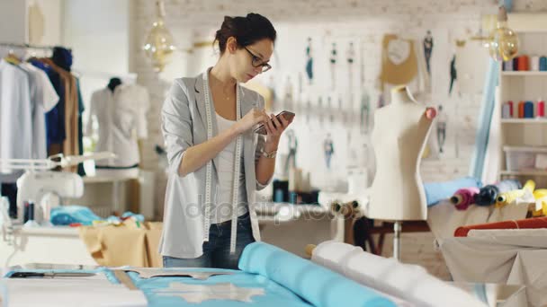 Hermosa morena diseñadora de moda establece plantillas en las telas y consulta su teléfono inteligente. Trabaja en un estudio de colores claros lleno de varias ropas, telas y bocetos en la pared . — Vídeos de Stock