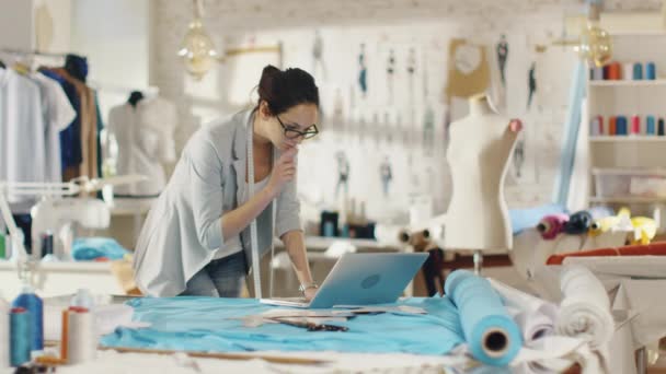 Hermosa diseñadora femenina utiliza el ordenador portátil en su escritorio que está cubierto con varias telas y artículos de costura. Su estudio es soleado, hay maniquí, ropa que cuelga y bocetos fijados a la pared . — Vídeos de Stock