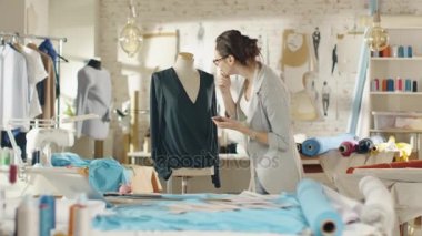 Güzel kadın moda tasarımcısı bluz bir manken olarak ayarlar, onun Smartphone ve gülümsüyor kullanır. Onun stüdyom Bright ve Sunny, Giysi asılı, renkli kumaşlar yalan masada.