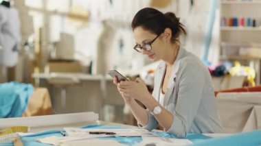 Güzel moda tasarımcısı masasında oturur, Smartphone kullanır. Onun stüdyom Sunny, tam renkli detaylarını, kumaşlar, dikiş öğeleri, skeçler.