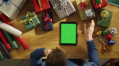 Top onun masada oturan bir genç adam görünümünü paketlenmiş hediyeler Green'de arıyorsunuz tam ekranlı Tablet bilgisayar. Sıcak güneş ışığı onun tabloda yer almaktadır..