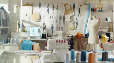 Bir kadeh bir güneşli moda tasarım stüdyosu. Biz kişisel bilgisayar, Giysi asılı, makine dikiş çalışma görmek ve çeşitli ilgili dikiş ayakta, renkli kumaşları mankenler masaya öğeleri.