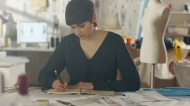 Güzel kadın moda tasarımcısı sanatçı onun Resepsiyon çizim çizimler yeni giyim koleksiyonu için oturuyor. Onun parlak ve Sunny stüdyosudur. Gördüğümüz çeşitli dikiş öğeleri, makineleri, skeçler, renkli kumaşlar rulo.