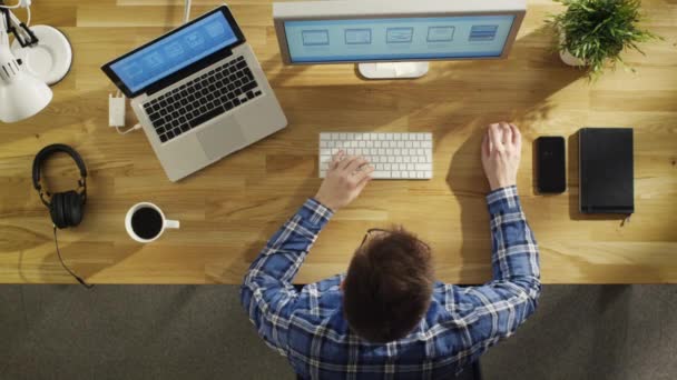 Bovenaanzicht van een jonge softwareingenieur bezig met zijn PC en Laptop. Geschoten op een mooie zomer-ochtend, valt zonlicht op zijn houten tafel. — Stockvideo