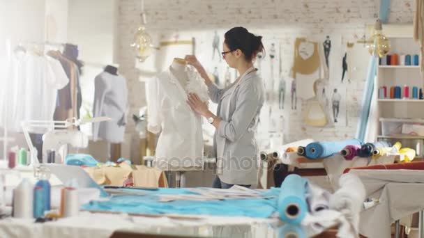 Mooie vrouw modeontwerper werken met afstemming Mannequin, Ornament vastzetten op een Shirt. Haar atelier is Bright en Sunny Mannequins staan rond, kleren opknoping, kleurrijke stoffen liggend op de tafel. — Stockvideo