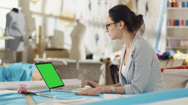 Schöne Frau Modedesignerin sitzt an ihrem Schreibtisch und arbeitet an Laptop mit grünem Bildschirm. ihr Atelier ist sonnig, bunte Stoffe und Schaufensterpuppen sind da. — Stockvideo