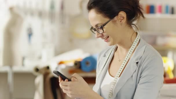 Die schöne Modedesignerin sitzt an ihrem Schreibtisch, tippt auf dem Smartphone. Ihr Atelier ist sonnig, voller farbenfroher Details, Stoffe, Nähsachen, Skizzen. — Stockvideo