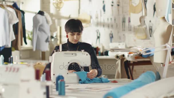 Vrouwelijke mode-ontwerper / naaister / naaister werkt op een naaimachine in haar zonnige Studio. Diverse naai artikelen en stoffen leggen rond Hare, zichtbaar schetsen aan de muur vastgemaakt. — Stockvideo