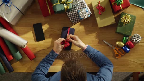 Vue du dessus du jeune homme emballant habilement des cadeaux colorés dans son studio ensoleillé. Smartphone avec écran vert maquillé repose sur la table . — Video