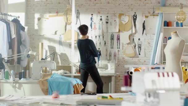 Kvinnliga modedesigner tittar på ritningar och skisser som är fästa på väggen. Studio är Sunny. Färgglada tyger, kläder hängande och sömnad objekt är synligt. — Stockvideo