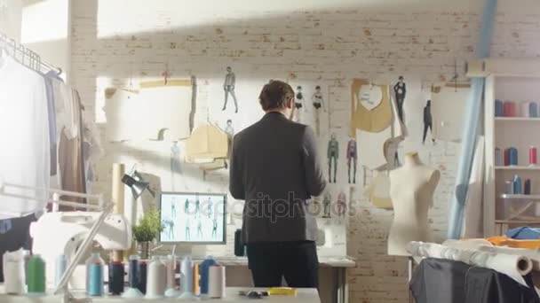 Mannelijke modeontwerper pinnen kleding schetsen aan de muur. Zijn atelier is Sunny, Personal Computer toont zijn werk. Kleurrijke stoffen, kleren opknoping en naaien Items zijn zichtbaar. — Stockvideo