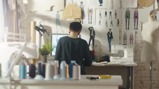 Vista posterior de una diseñadora de moda femenina que trabaja en una computadora personal, tomando notas. Su lugar de trabajo es soleado, máquina de coser de pie sobre su mesa, bocetos de la futura colección fijada a la pared . — Vídeos de Stock