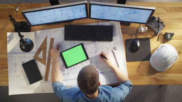 Vista superior del ingeniero arquitectónico se basa en sus planos, se compara con la computadora de la tableta con la pantalla verde, usando la computadora de escritorio también. Su escritorio está lleno de objetos útiles y sol de noche . — Vídeos de Stock