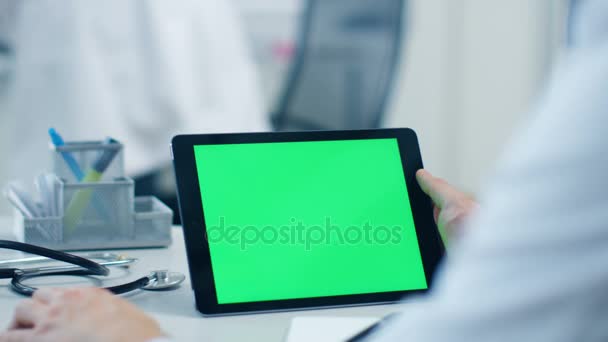 Närbild av en manlig läkare använder Tablet PC med grön skärm, hans assistent arbetar i bakgrunden. Office är ljust, vitt och modernt. — Stockvideo