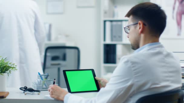 Close-up of a Male Doctor Uses Tablet Computer with Green Screen, He also Holds Pen in his Notepad, His Assistant Works in the Background. La oficina es brillante, blanca y moderna . — Vídeos de Stock