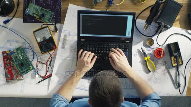 Vista dall'alto di un tecnico IT dotato che scrive codice sul suo computer portatile seduto alla sua scrivania, è circondato da vari componenti tecnici. Sole splende sulla sua scrivania . — Video Stock