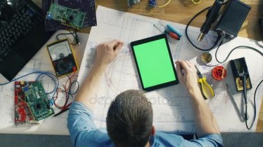 Üst görüntüleme erkeği olarak bu planları üzerinde çalışan teknisyeni, bir yeşil ekran Tablet bilgisayarda sallıyorsun. Surround devre kartları, dizüstü bilgisayar ve diğer teknik bileşenleri tarafından o. Onun masada güneş parlar.