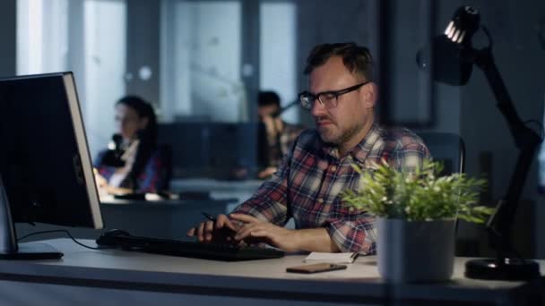 Késő este az irodában. Ember ül az íróasztalánál, szerkezet-ra egy személyes számítógép, írja le valamit a Jegyzettömb. Munkatársaimmal látható a háttérben. — Stock videók