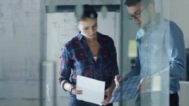 Gece geç saatlerde erkek ve kadın güneş mühendisleri Taslaklar ve bir güneş paneli parçası tartışmak. Kadın Taslaklar, adam tutun Panel tutar. Arka plan beyaz tahta'yı skeçler, duvar Blueprints Pinned ile.