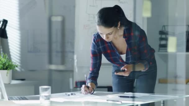 Engenheira de design feminina trabalha em documentos em uma sala de conferências, check-up de última hora, usa seu smartphone. No quadro branco de fundo com esquemas nele, vários planos pendurados nas paredes . — Vídeo de Stock