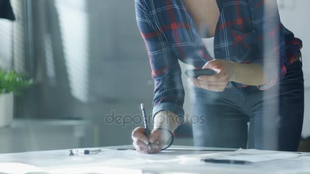 Close-up of a Female Design Engineer Works on Documents in a Conference Room, Last Minute Check-up, Uses Her Smartphone. In the Background Whiteboard with Schemes on it, Various Blueprints Hanging on the Walls. — Stock Video