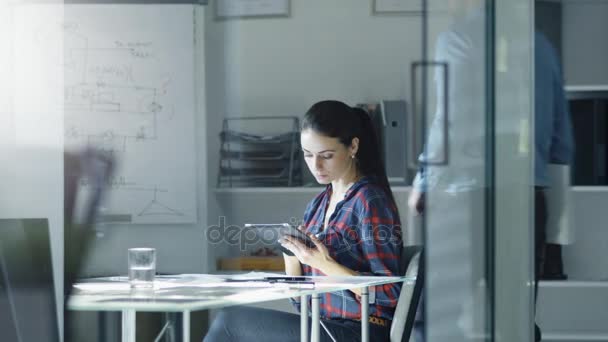 Engenheira de design feminina senta-se na mesa de vidro em seu escritório, trabalha em um computador tablet, Blueprints Deitado em sua mesa. Vem aí o Colega. No quadro branco de fundo com diagramas e esboços . — Vídeo de Stock