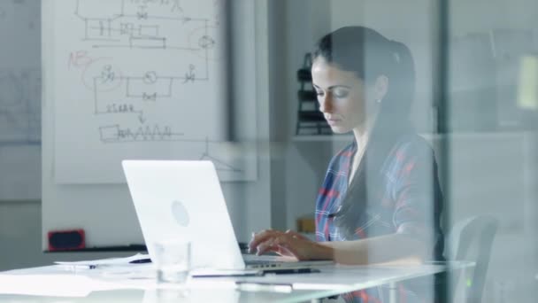 Ingeniera de diseño femenina se sienta en la mesa de vidrio de su oficina, tipos en un ordenador portátil, planos colocados en su escritorio. En la pizarra de fondo con diagramas y bocetos . — Vídeos de Stock