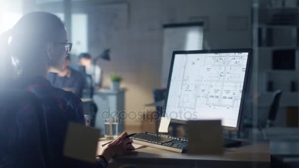 Tarde en la noche en la Oficina de Ingeniería Diseñadora femenina trabaja en una computadora personal que muestra planos de su proyecto. En el fondo, sus colegas trabajando . — Vídeos de Stock