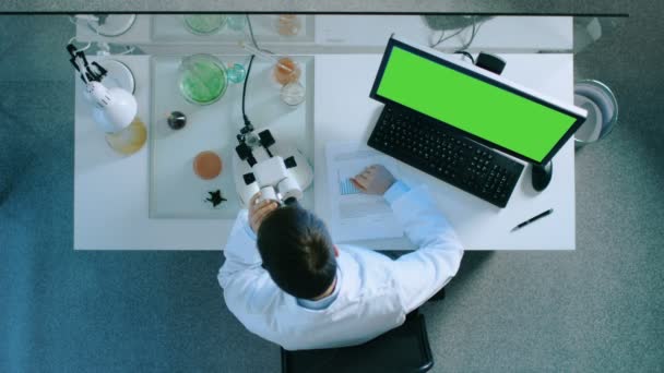 Draufsicht eines Wissenschaftlers, der an einem PC mit grünem Bildschirm arbeitet, ins Mikroskop schaut und die Ergebnisse auf ein Dokument mit Diagrammen schreibt. — Stockvideo