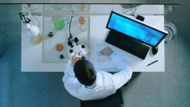 Vista dall'alto di uno scienziato che guarda al microscopio e analizza le informazioni con il computer . — Video Stock