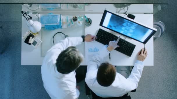 Draufsicht auf zwei Ärzte am Arbeitstisch, die das Röntgenbild des Patienten diskutieren, das auf einem Monitor gezeigt wird. sie kommen zu dem Schluss und die Ärzte schreiben die Diagnose auf einen Zettel. — Stockvideo
