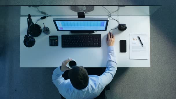 Bovenaanzicht van een kantoor werknemer zit aan zijn bureau werken met werkbladen op zijn PC. It's Late avond en hij drinkt koffie. — Stockvideo