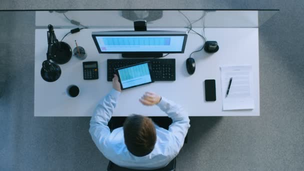 Ein Büroangestellter sitzt an seinem Schreibtisch und blättert auf seinem Tablet-Computer in Tabellen. er benutzt auch seinen PC. — Stockvideo