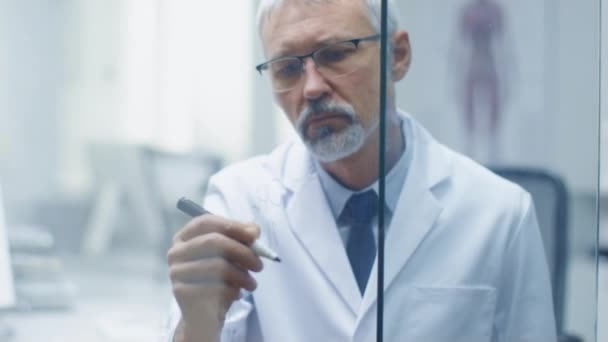 Un chercheur scientifique chevronné écrit une formule scientifique sur un tableau blanc en verre. Il porte des lunettes et son bureau est lumineux et moderne . — Video