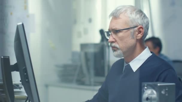 Engineering-Bureau. Chief Engineer werken achter zijn bureau, sommige technische onderdelen liggend op zijn bureau. Zijn assistent werkt aan zijn Bureau op de achtergrond. — Stockvideo