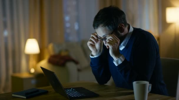 Cansado hombre de mediana edad trabaja en un ordenador portátil. Tiene los ojos fatigados, temporalmente se quita los anteojos y se frota los ojos. Su apartamento se ve acogedor en tonos amarillos . — Vídeos de Stock