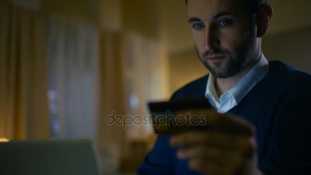 Gros plan d'un homme assis à son bureau avec un ordinateur portable détenant une carte de crédit et faisant un achat sur Internet. Son appartement a des connotations jaunes et se réchauffe . — Video