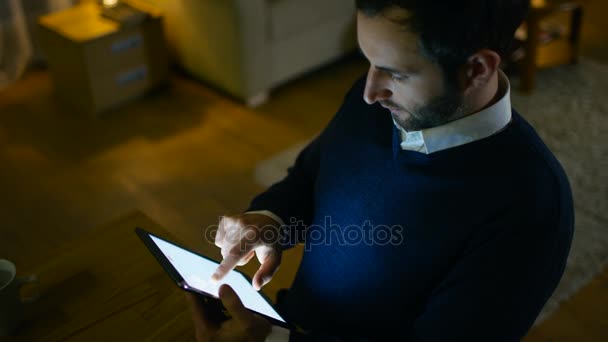 Ein respektabler Mann mittleren Alters sitzt an seinem Schreibtisch und benutzt einen Tablet-Computer. Er ist allein zu Hause. im Hintergrund sieht seine Wohnung in gelben Farben stylisch aus. — Stockvideo