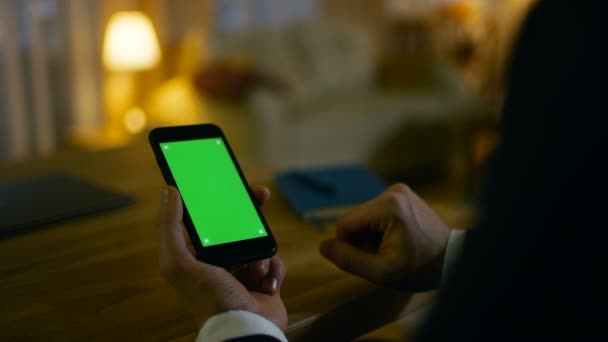 At Home Man Senta-se em sua mesa e usa Smartphone com tela verde nele. Ele bebe de uma taça. Seu apartamento é feito em cores amarelas e é quente . — Vídeo de Stock