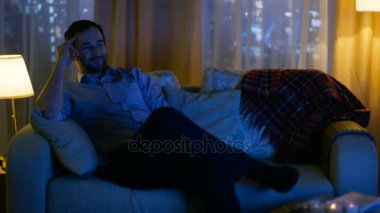 Akşam orta yaşlı adam evde rahat bir kanepede oturan ve Tv izliyor. Eğlenin görünüyor. Büyük şehir manzaralıdır gökdelenler ile onun Windows.