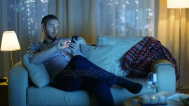 Akşam orta yaşlı adam oturur bir divan Smartphone kullanır. TV fiske üzerinde döndü, onu aydınlatır. Büyük bir şehirde gökdelenler ile arka plan Windows görünüm.