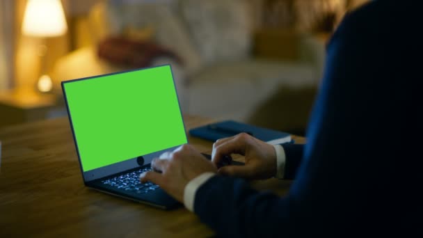 At Home Man Sits at His Desk and Types on a Laptop with Green Screen on It. Su apartamento está hecho en colores amarillos y es cálido . — Vídeos de Stock