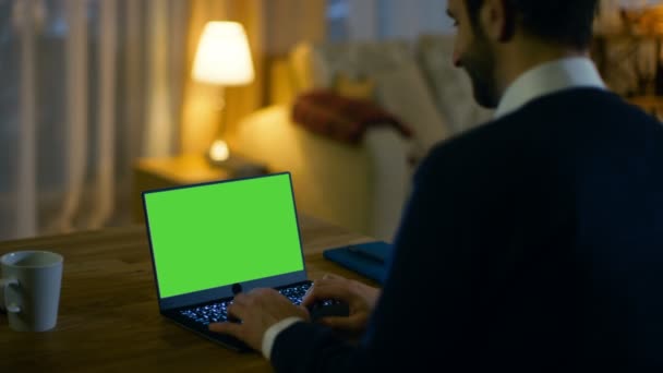 En casa Vista trasera de un hombre sentado en su escritorio y escribiendo en un ordenador portátil con pantalla verde en él. Su apartamento está hecho en colores amarillos y es cálido . — Vídeos de Stock