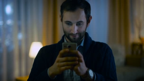 Retrato de un hombre en casa sosteniendo su teléfono inteligente con ambas manos. Su apartamento está hecho en colores amarillos cálidos y tiene vista a rascacielos. Las luces están encendidas. . — Vídeo de stock