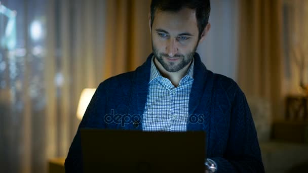 Retrato de un hombre en casa trabajando en una computadora. La pantalla lo ilumina. Su apartamento está hecho en colores amarillos cálidos y tiene vista a rascacielos . — Vídeos de Stock