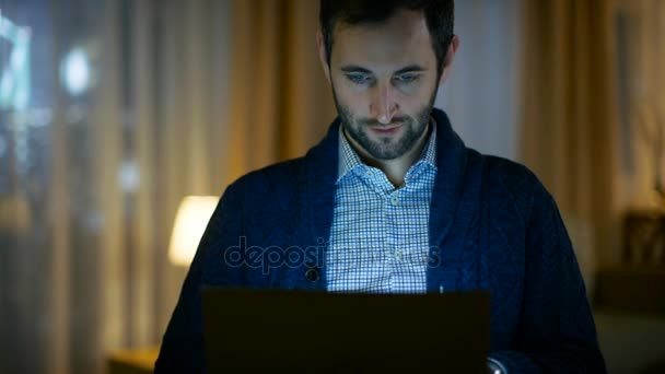 Retrato de un hombre en casa trabajando en una computadora. La pantalla lo ilumina. Su apartamento está hecho en colores amarillos cálidos y tiene vista a rascacielos . — Vídeo de stock