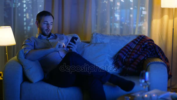 In the Evening Middle Aged Man Sits on a Sofa Uses Smartphone. Encendió la televisión lo ilumina. Vista de fondo de una gran ciudad con rascacielos . — Vídeo de stock